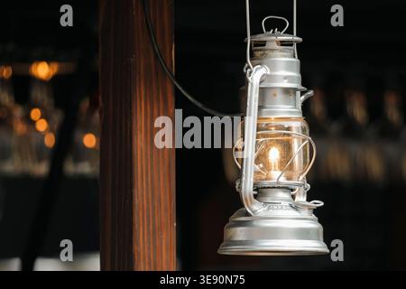 Vintage metal lantern with clear glass and a warm glowing bulb hangs from a wooden post, creating a cozy rustic atmosphere. Stock Photo