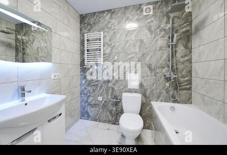 Bright bathroom interior with bathtub, sink vanity and toilet in gray stone finish. Contemporary washroom with rainfall shower, heated towel rail, lighted mirror and sleek design Stock Photo