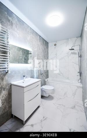 Contemporary bathroom interior with bathtub, suspended toilet and sink cabinet. Refined washroom with cool gray stone texture, white marble wall panels, backlit mirror and towel radiator Stock Photo
