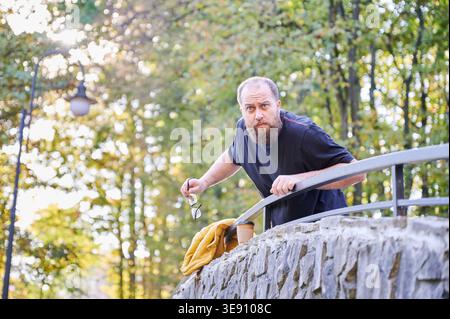 Bearded man searching for keys on stone bridge, peering over railing, holding small keyring, yellow jacket draped over wall, concerned expression, late afternoon light through trees Stock Photo