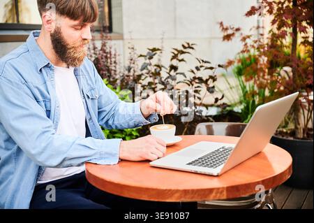 Casual remote worker sits at patio table with laptop and cappuccino, stirring beverage in calm courtyard setting with greenery and contemporary wall Stock Photo