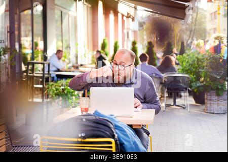Man eating meal at outdoor cafe while working on laptop, sitting among other visitors, focused on screen, combining lunch and remote work in busy urban environment Stock Photo