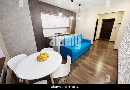 Modern studio apartment with blue sofa, dining table and compact kitchen in open plan interior. Stylish home design with wooden floor, pendant lights and urban living concept Stock Photo