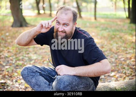 Man points to temple and smiles while holding eyeglasses in autumn park. Friendly outdoor portrait for idea, intelligence, smart thinking, creativity, or lifestyle promotion Stock Photo