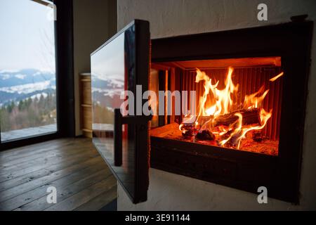 Fire burns in modern fireplace inside cozy home. Close up of glowing logs and flames, glass door open, wooden floor and mountain view through window, warmth and comfort concept Stock Photo
