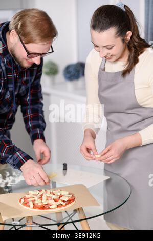 Happy young couple smiling, adding cheese toppings to homemade pizza in modern kitchen. Man and woman cooking together at home, culinary hobby, teamwork, romantic relationship concepts, family bonding Stock Photo