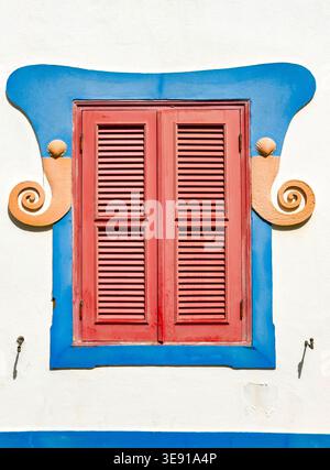 Red louvered window shutters with blue decorative frame and shell ornaments on a white wall in lagos algarve Portugal Stock Photo