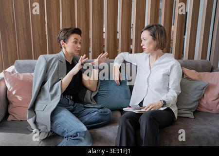 Female colleagues gesturing and chatting while sitting together on sofa in modern office. Chatting, gesture, colleagues, teamwork, office. Stock Photo