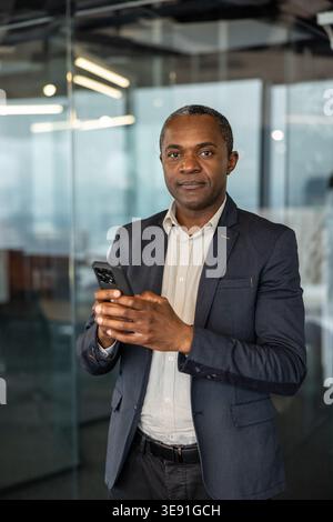 Confident professional businessman standing in a modern office, holding a mobile phone, representing communication, leadership, and digital technology in the workplace Stock Photo