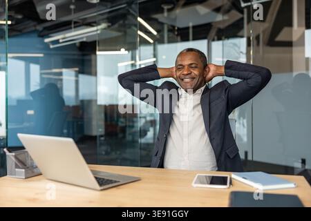 Mature man in a suit relaxing and stretching with hands behind his head at a modern office desk, smiling with eyes closed, enjoying a successful break after finishing tasks Stock Photo