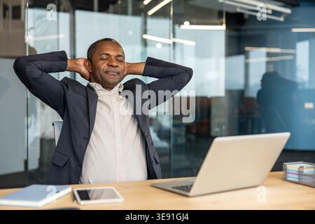 Mature professional man taking a moment to unwind, leaning back with hands behind head and eyes closed at his desk with a laptop, finding comfort and satisfaction in a successful day Stock Photo