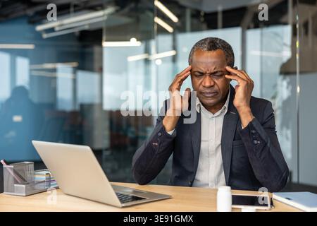 Mature male entrepreneur suffering from an intense headache at his modern office desk, pressing temples with hands, experiencing stress, burnout, and frustration from work pressure Stock Photo