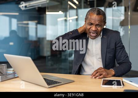 Mature man working on a laptop experiencing discomfort and strain in his neck, holding it with his hand while feeling pain from poor posture or prolonged computer use in an office setting Stock Photo