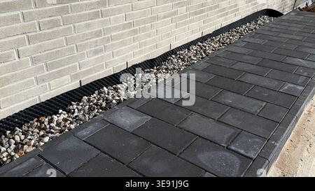 Paved walkway made of dark gray concrete blocks alongside a textured brick wall with gravel and drainage visible in the landscaping Stock Photo