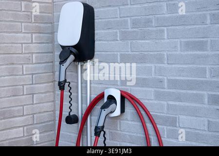 Electric vehicle charging station mounted on a textured brick wall, featuring two charging connectors with red cables coiled and ready for use Stock Photo