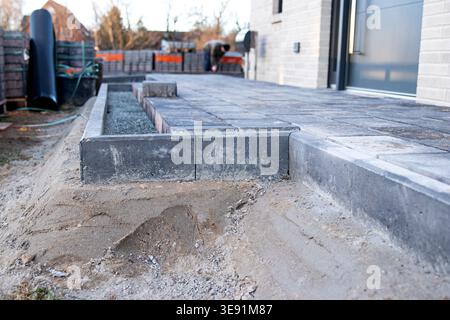 Paved outdoor patio area under construction with gray stone blocks arranged neatly, sand and gravel visible, and a modern house in the background Stock Photo