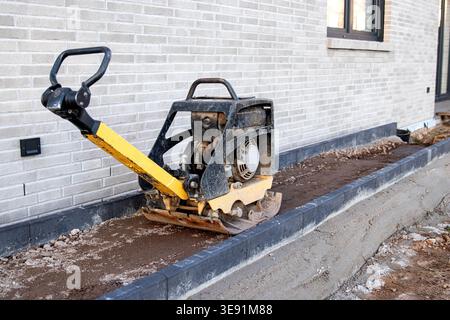 Compactor machine with yellow handle on freshly leveled soil near a brick wall of a modern building under construction in a residential area Stock Photo