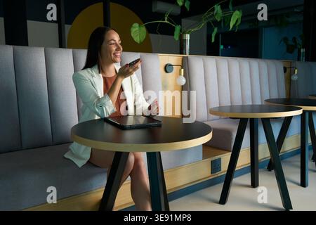 Happy businesswoman talking on speaker smartphone while sitting alone in office lobby Stock Photo