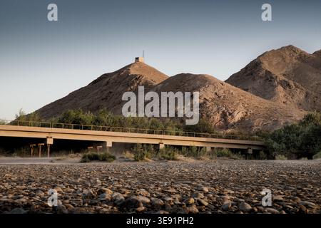A bridge crosses a dry riverbed beneath rocky hills under soft evening light. Stock Photo