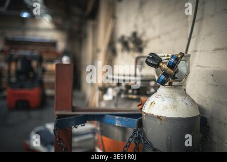 Close-up of an industrial gas cylinder with a valve. The cylinder is placed in a workshop with a forklift truck in the blurred background. Stock Photo