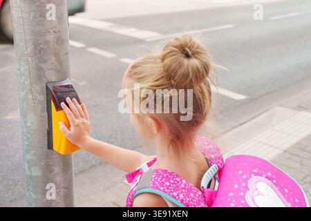 Schoolgirl with schoolbag presses yellow button pedestrian crossing to turn on green light traffic light to cross street of city. Concept of primary s Stock Photo