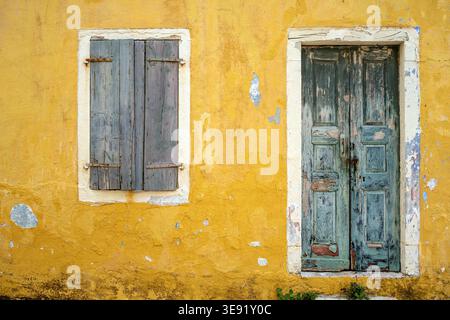 Weathered blue wooden door and closed shutters on a yellow stucco wall with peeling paint and cracks, showing aged Mediterranean architecture. Stock Photo