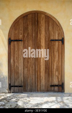 Arched wooden double door with vertical planks and iron hinges set in a stucco wall, showing weathered texture and rustic architectural detail. Stock Photo