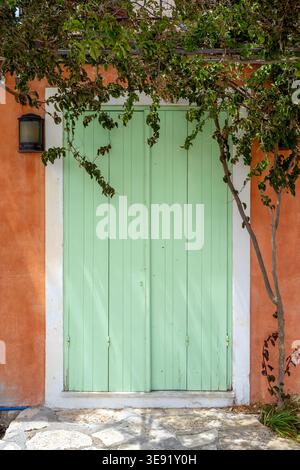 Mint green wooden double door with vertical panels set in an orange stucco wall, framed by foliage and branches. Stock Photo