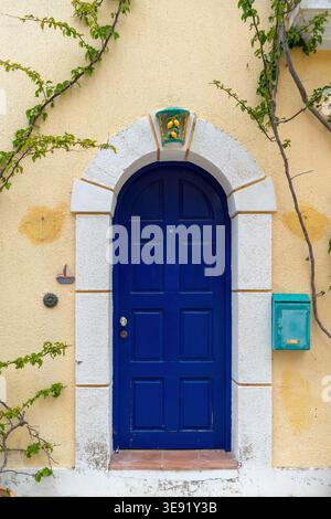 Blue arched wooden door with decorative panels set in a yellow stucco wall, framed by white stone and climbing vines. Stock Photo
