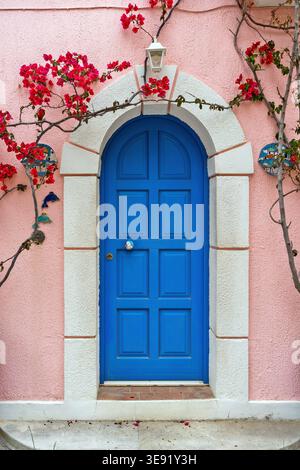 Bright blue arched wooden door set in a pink stucco wall with white stone frame, decorated with bougainvillea flowers and ceramic ornaments. Stock Photo