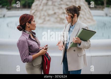 Two businesswomen discussing documents outdoors by a fountain during a professional conversation Stock Photo