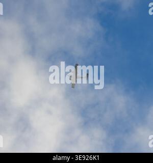 Small airplane flying through partly cloudy blue sky. Wings visible as it moves across bright sky. Sky filled with white clouds and blue patches. Clea Stock Photo
