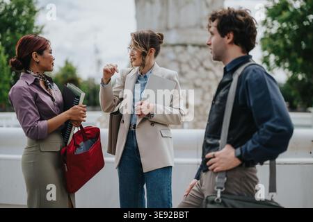 Three colleagues having a casual business conversation outdoors near a fountain while holding laptops and bags Stock Photo
