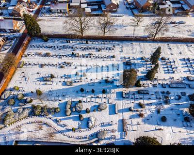Aerial top-down view of a peaceful cemetery covered in fresh white snow. Rows of headstones and winter trees create long shadows in the bright morning Stock Photo