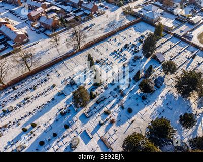 Aerial top-down view of a peaceful cemetery covered in fresh white snow. Rows of headstones and winter trees create long shadows in the bright morning Stock Photo