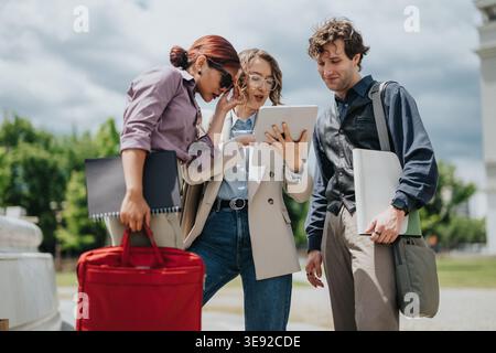 Three young business colleagues collaborating outdoors while reviewing a tablet during a casual urban meeting Stock Photo