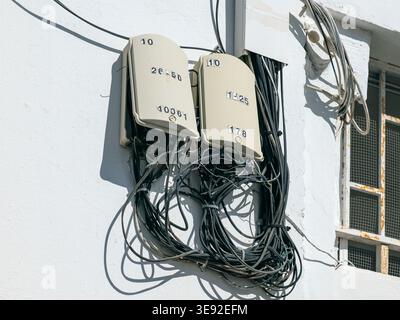 Two beige telecommunications junction boxes mounted on a white wall with numerous black cables emerging and hanging below, showing a complex network. Stock Photo