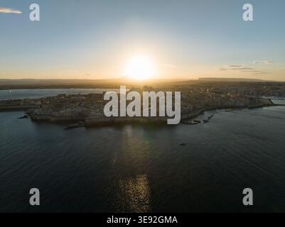 Aerial view of the historic Ortigia island at sunset with golden light reflecting on the sea and ancient architecture in Syracuse, Sicily, Italy. Stock Photo