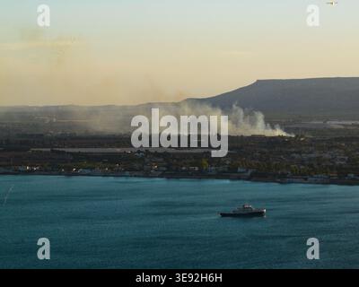 Aerial view of smoke rising from a coastal landscape with a boat on the water and hills in the distance in Syracuse, Sicily, Italy. Stock Photo