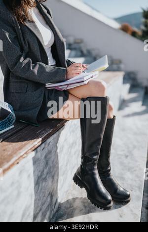 Student writing notes outdoors on bench in stylish boots and blazer during study break Stock Photo