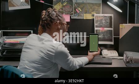 Law officer holding mobile pohne with greenscreen, young detective looking at blank copyspace display. Female detective in incident room using chroma key mockup template in archive. Stock Photo
