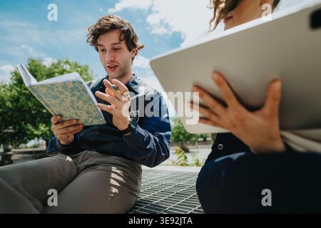 Business coworkers collaborating outdoors while reading a notebook and using a tablet during a casual meeting Stock Photo