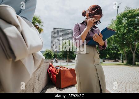 Businesswoman adjusting sunglasses while checking documents outdoors with coworkers in urban park Stock Photo