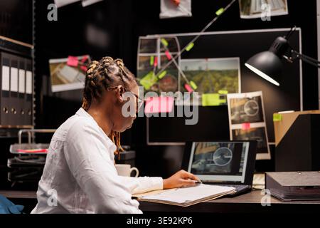 African american woman private detective studying clues in agency office. Police investigator sitting at desk, looking at suspect photo on laptop and reading criminal case information Stock Photo