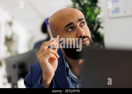 Arab entrepreneur thinking and looking away while analyzing sales strategy on laptop. Young company employee managing financial data while working in start up business office Stock Photo
