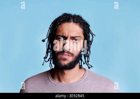 Arab confused man frowning eyebrows and looking at camera. Young adult arabian brunette person with braided hairstyle with unsure facial expression close up studio portrait Stock Photo