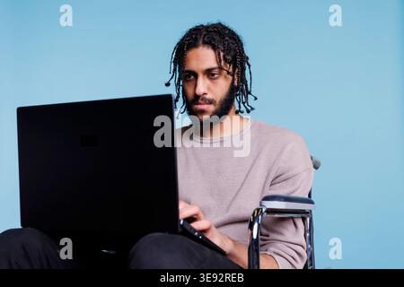Focused programmer in wheelchair developing software and typing on laptop. Concentrated arab person with disability reading report and planning work using portable computer Stock Photo