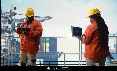 Engineer on offshore platform deck using artificial intelligence software on laptop to optimize drilling operations. Teamworking colleagues monitoring barge activity using AI technology, Stock Photo