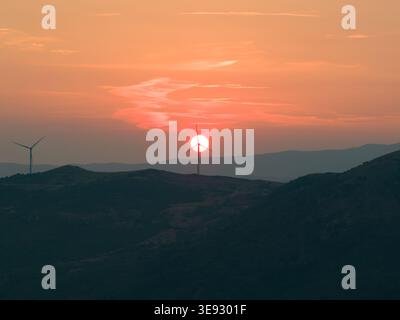 Aerial view of wind turbines on rolling hills with the sun setting behind a turbine on the horizon in Castel di Lucio, Sicily, Italy. Stock Photo