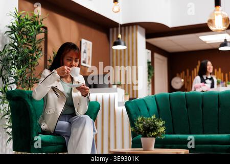 Asian tourist enjoying coffee in hotel lobby. Customer holding a cup of coffe in luxury hotel. Female client drinking tea and relaxing in hotel lounge. Hotel guest having a drink. Stock Photo